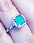 A natural Colombian emerald cushion cut with a halo diamond in white gold on a finger.