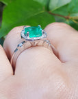On top of a finger wearing a white gold cushion cut emerald ring on a leafy background.