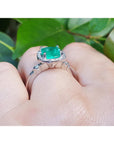 On top of a finger wearing a white gold cushion cut emerald ring on a leafy background.