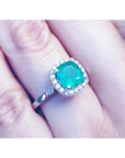 A natural Colombian emerald cushion cut with a halo diamond in white gold on a finger.