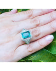 Hand wearing an emerald ring with side trillion cut diamonds against a leafy background.