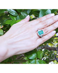 Hand wearing a Ladies' natural Colombian emerald ring crafted in 18k white gold against a green leafy background.