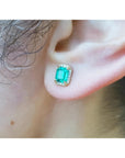 An ear wearing a natural emerald stud earring with halo diamond in 14k gold.