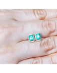 Emerald green gemstone earrings held between fingers on a light skin background.