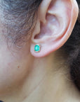 Close-up of a woman's ear wearing a yellow gold emerald stud earring with blurred background.