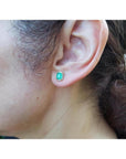 Rectangular Colombian emerald halo stud earrings in 14k yellow gold, worn on an ear.