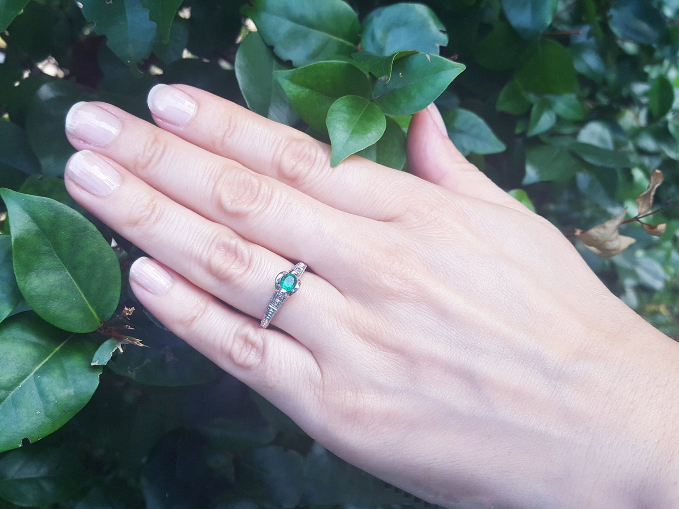 Hand wearing a natural oval cut colombian emerald ring against a leafy background.