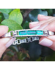 Emerald and princess cut diamond bracelet held against green leaves.