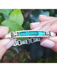 Emerald and princess cut diamond bracelet held against green leaves.