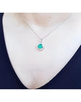 Cushion-cut Colombian emerald pendant with diamond halo on a white gold chain, worn on a dark top.