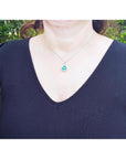 A woman wearing a black top with a necklace featuring a natural emerald pendant against a blurred green background.