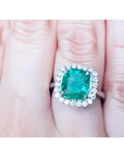 An emerald engagement ring cushion cut with a halo of natural diamond in white gold on a finger.