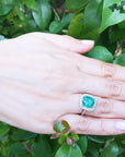 Hand wearing a cushion cut Colombian emerald ring against a green leafy background.