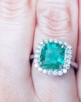 An emerald engagement ring cushion cut with a halo diamond on a finger.