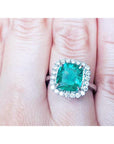 An emerald engagement ring cushion cut with a halo diamond on a finger.