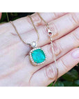 Cushion cut emerald necklace with halo diamonds on an 18K yellow gold chain, held in a hand.