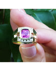 Gold ring with a pink natural sapphire gemstone held between fingers against a green leafy background