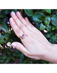 Hand wearing a gold ring with a pink natural sapphire gemstone against a green leafy background