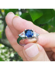 White gold ring with a natural blue sapphire cushion cut held in a hand against a blurred green background