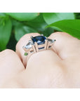 Cushion cut deep blue sapphire ring with trillion side sapphires in 14K white gold on a hand.