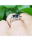 Cushion cut deep blue sapphire ring with trillion side sapphires in 14K white gold on a hand.