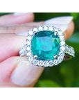 Emerald ring with diamond halo held between fingers against a blurred green background.