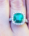 Cushion cut emerald ring with diamond halo on a finger.