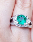 A cushion cut emerald engagement ring with diamond accents on a finger.