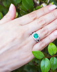 Hand wearing an emerald ring with a green leafy background.