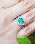 A white gold cushion cut emerald ring on a hand with a natural background.