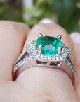 Emerald ring with diamond accents on a finger against a blurred green background.
