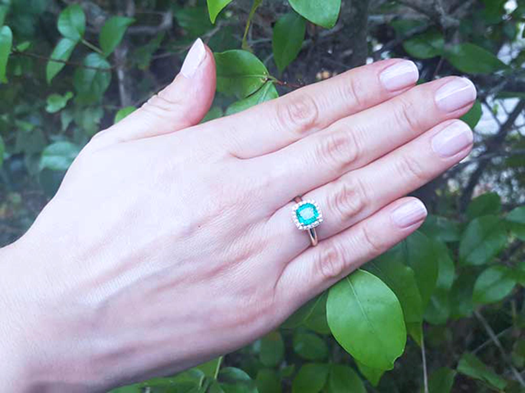 Hand wearing a cushion cut emerald ring with a leafy background.