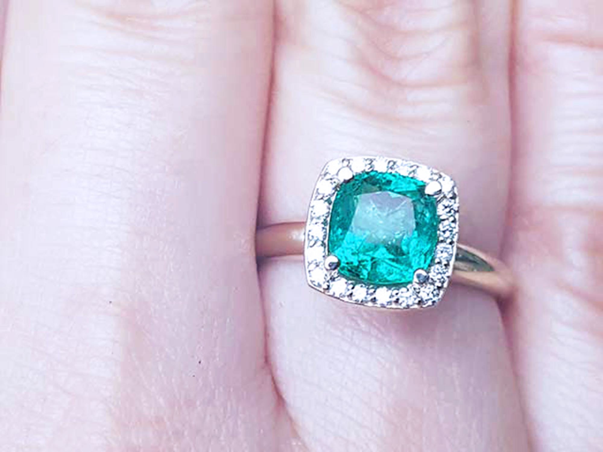A women's cushion cut emerald ring with halo diamond on a finger.