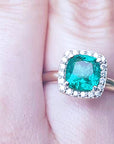 A women's cushion cut emerald ring with halo diamond on a finger.