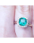 A women's cushion cut emerald ring with halo diamond in 14k yellow gold on a finger.