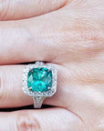 A natural cushion cut emerald engagement ring with halo diamond on a hand.