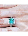 A natural cushion cut emerald engagement ring with halo diamond on a hand.