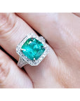 A lady's emerald ring cushion cut with a halo diamond crafted in 18k white gold on a finger.