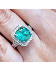 A lady's emerald ring cushion cut with a halo diamond crafted in 18k white gold on a finger.