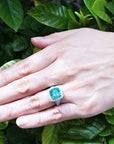 Hand wearing a ladies emerald cushion cut ring with a leafy background.