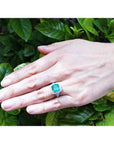 Cushion cut emerald engagement ring with diamond halo in 18K white gold on a hand.