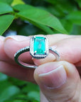 A natural emerald ring held between fingers against a blurred green leafy background.