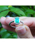 14K white gold cushion-cut emerald solitaire ring with rope detailing, held against green leaves.