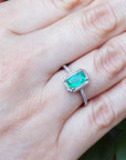 A cushion cut emerald ring on a hand with a green leaf background.