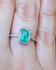 A cushion cut emerald ring on a finger with a blurred green leaf background.