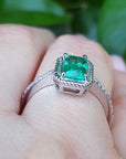 A white gold emerald ring on a hand against a green leafy background.