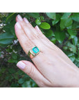 Hand wearing a gold men's ring with a cushion cut natural emerald against a green leafy background.