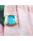 Gold ring for men with a cushion cut Muzo colombian emerald on a finger against a blurred natural background.