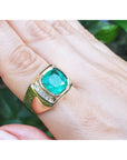 A 18k yellow gold mens emerald ring bezel set on a hand against a blurred green background.