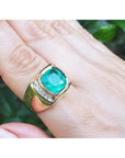 A 18k yellow gold mens emerald ring bezel set on a hand against a blurred green background.